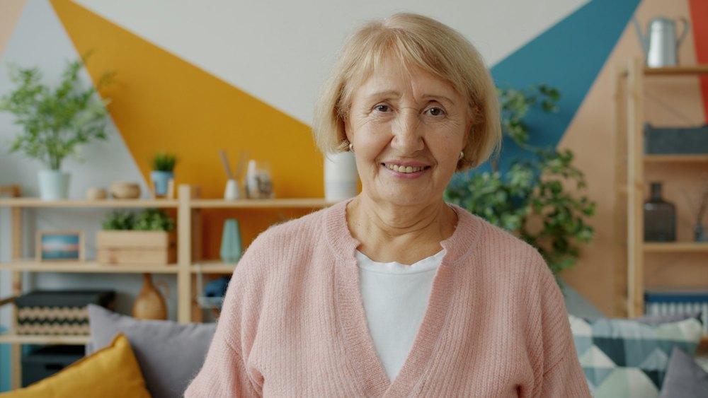 Lächelnde ältere Dame in rosa Strickjacke in ihrem Wohnzimmer